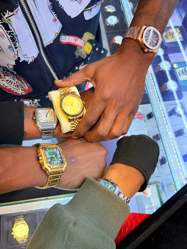 Three wrists over a lit counter, each trying on a watch: a gold one held on its cushion, a steel square one and a gold square one with a green dial
