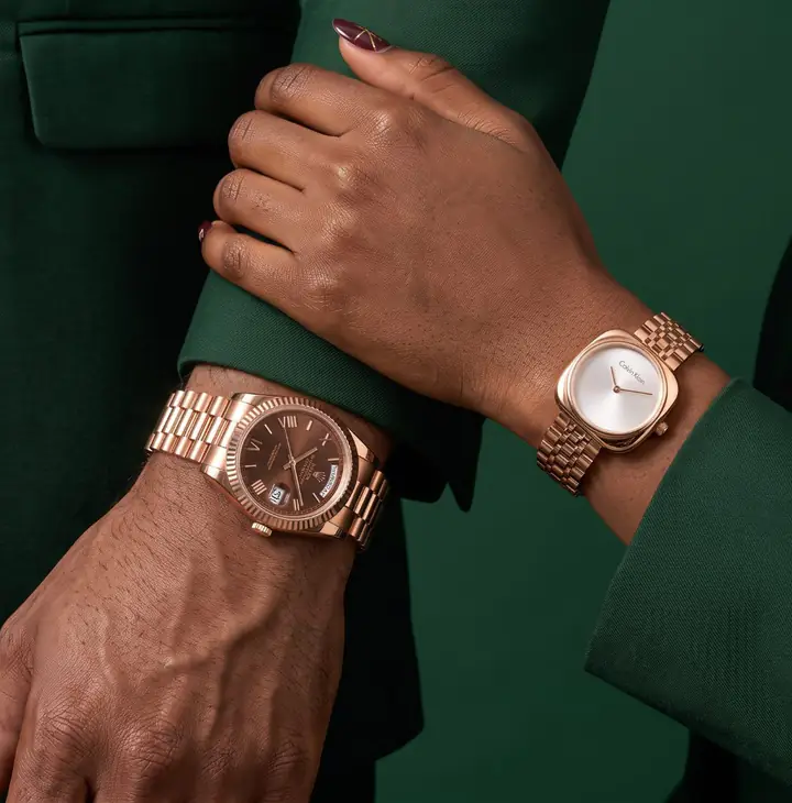 Two wrists in dark green suit sleeves: his rose-gold watch with a brown dial and fluted bezel, her rose-gold watch with a white cushion-shaped dial